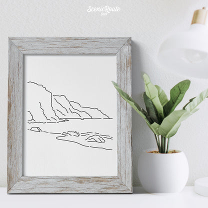 A framed line art drawing of The NaPali Coast on a desk with a plant and lamp