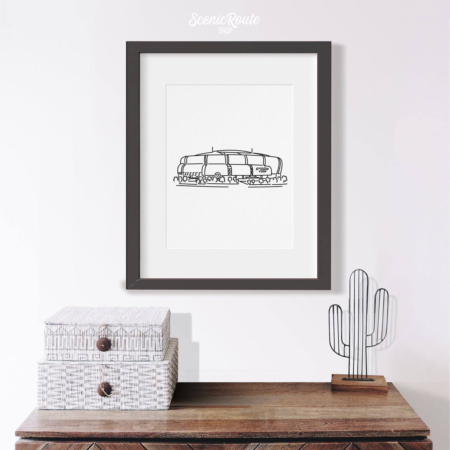 A framed line art drawing of the Cardinals Stadium on a wall above a small table with a saguaro figurine