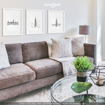 A group of three framed drawings on a white wall hanging above a couch with pillows and a blanket. The line art drawings include the New York Skyline, Chrysler Building, and Empire State Building