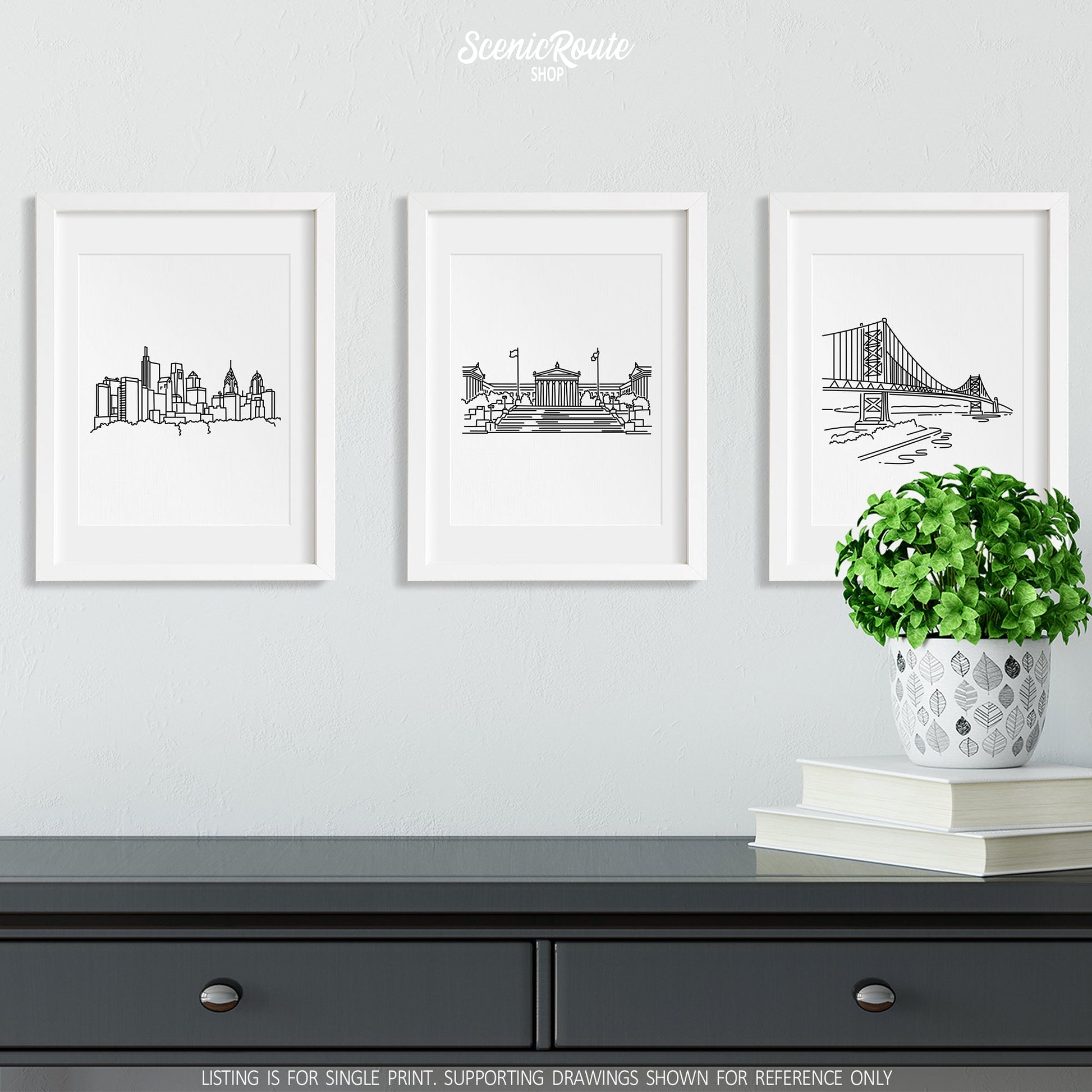 A group of three framed drawings on a wall above a dresser with books and a plant. The line art drawings include the Philadelphia Skyline, Philadelphia Art Museum, and Ben Franklin Bridge