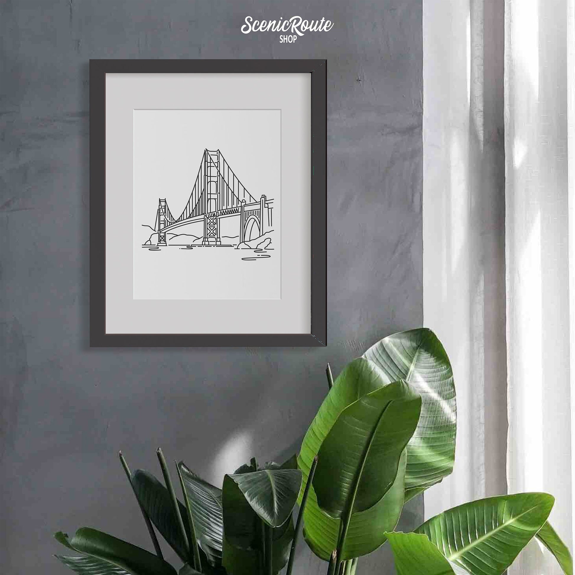 A framed line art drawing of the Golden Gate Bridge on a wall above a tropical plant