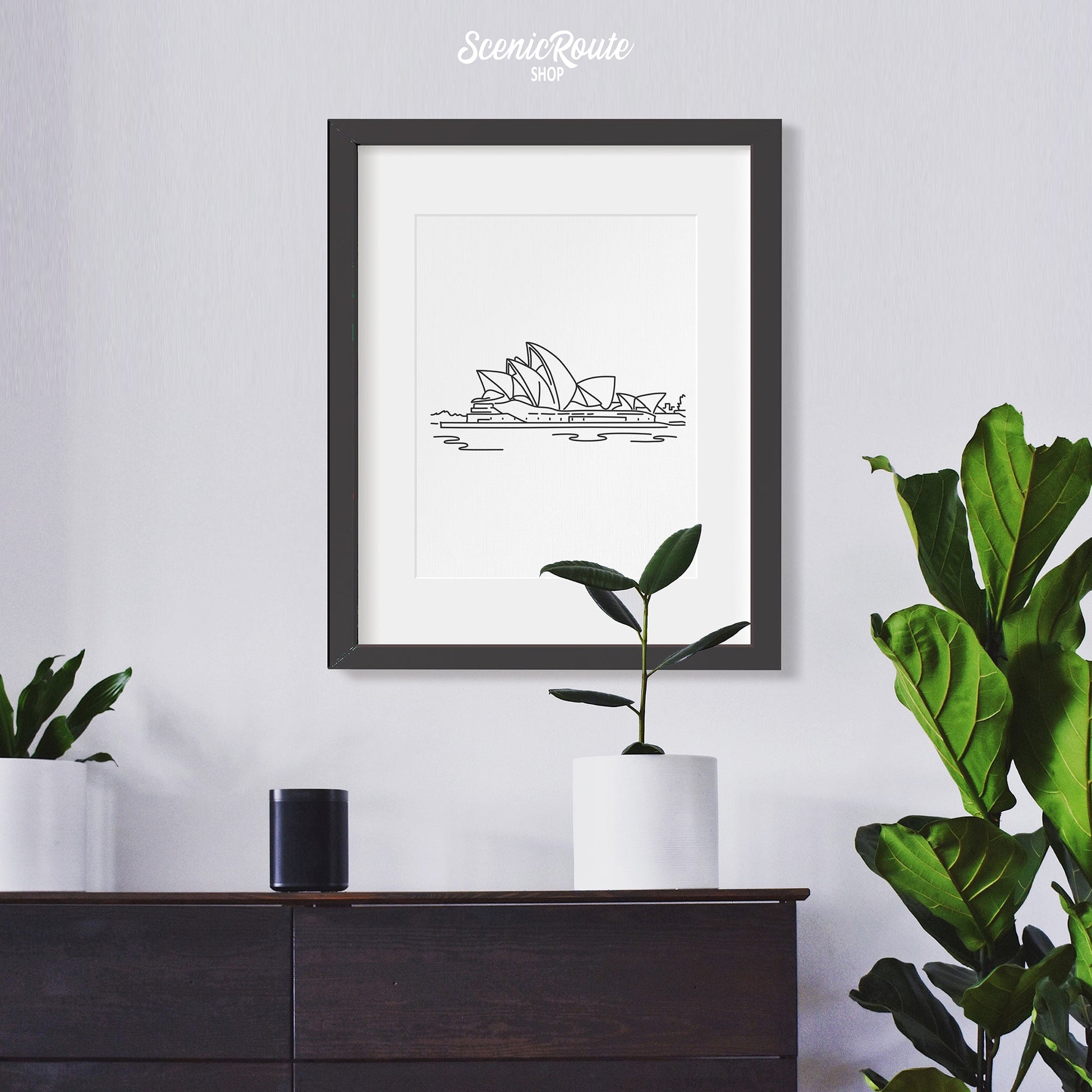A framed line art drawing of the Sydney Opera House above a dresser with plants