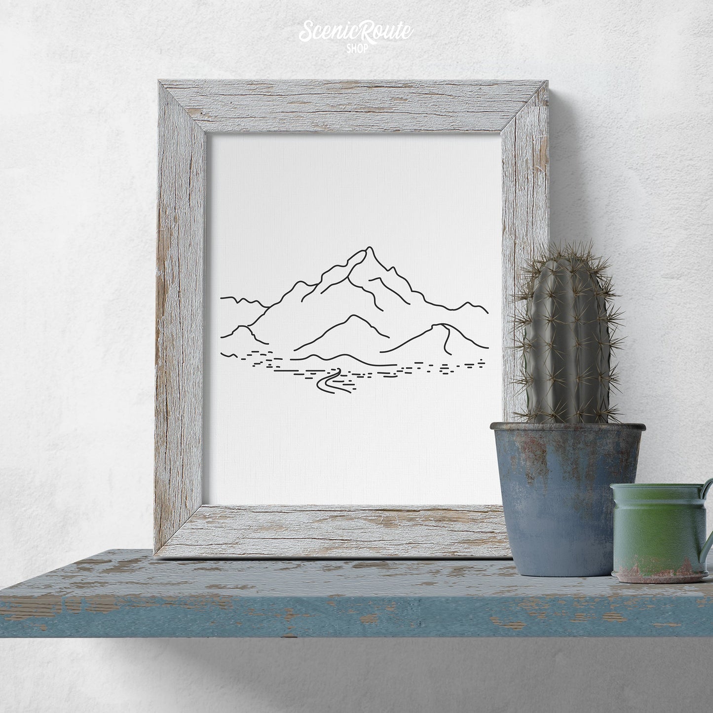 A framed line art drawing of Piestewa Peak on a blue shelf with a potted cactus