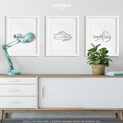 A group of three framed drawings on a credenza with a lamp and plant. The line art drawings include the Portland Skyline, Mount Hood, and Haystack Rock