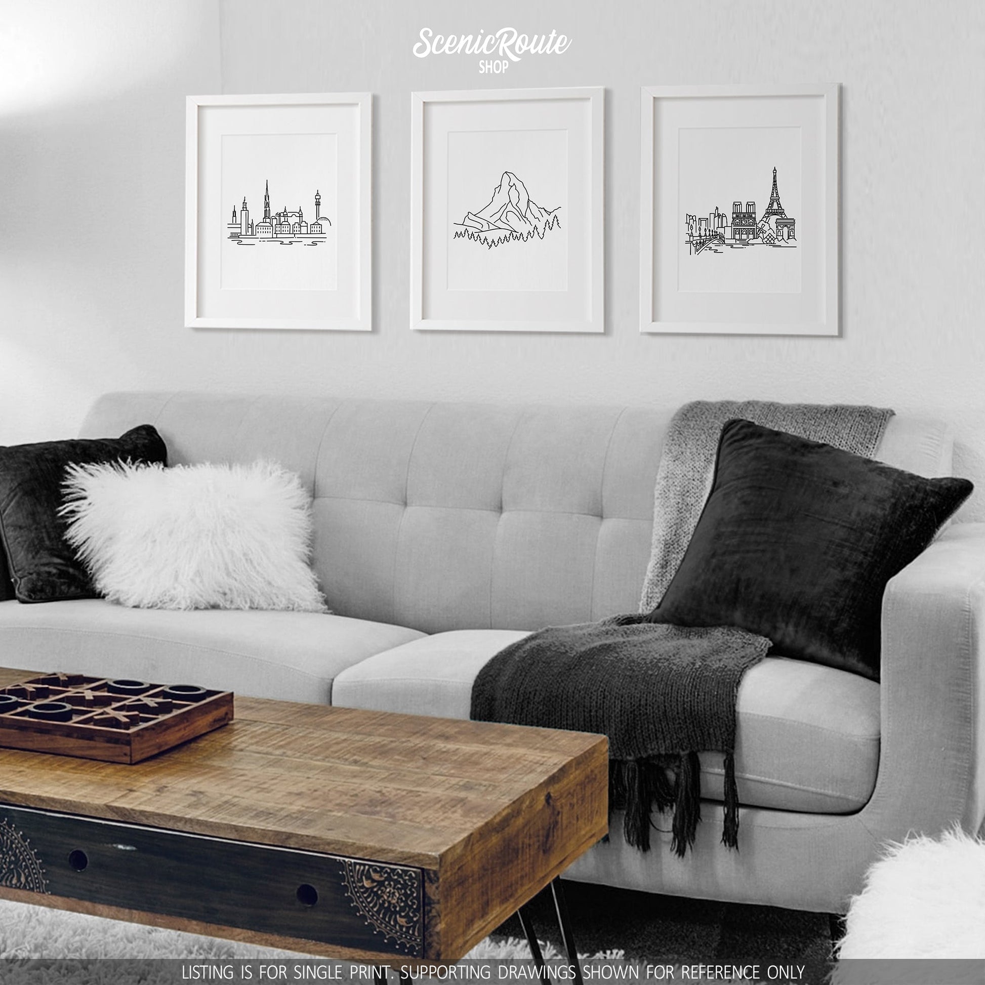 A group of three framed drawings on a white wall hanging above a couch with pillows and a blanket. The line art drawings include the Stockholm Skyline, the Matterhorn, and the Paris Skyline