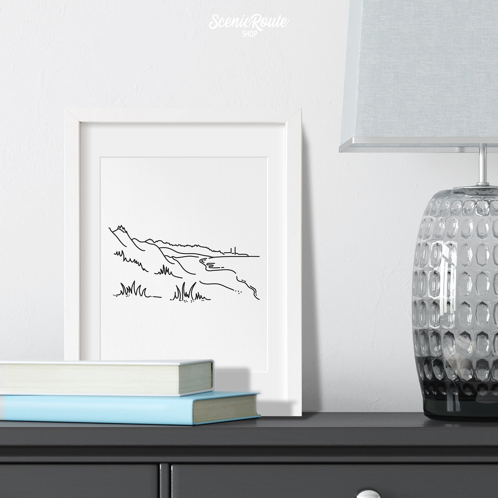 A framed line art drawing of Indiana Dunes National Park on a dresser with books and a lamp
