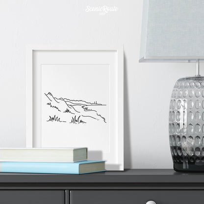 A framed line art drawing of Indiana Dunes National Park on a dresser with books and a lamp