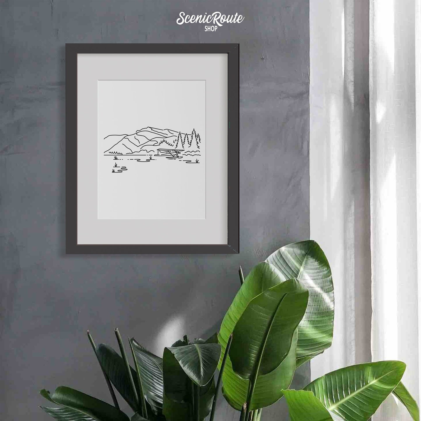 A framed line art drawing of Lake Clark National Park on a gray wall above a tropical plant