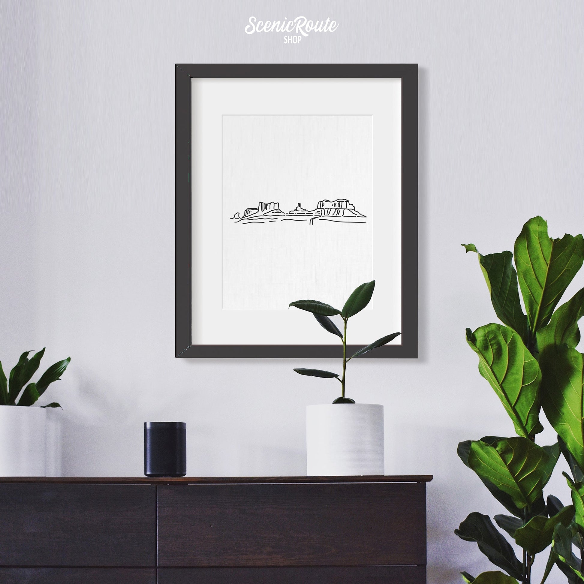 A framed line art drawing of Monument Valley hanging over a dresser with plants