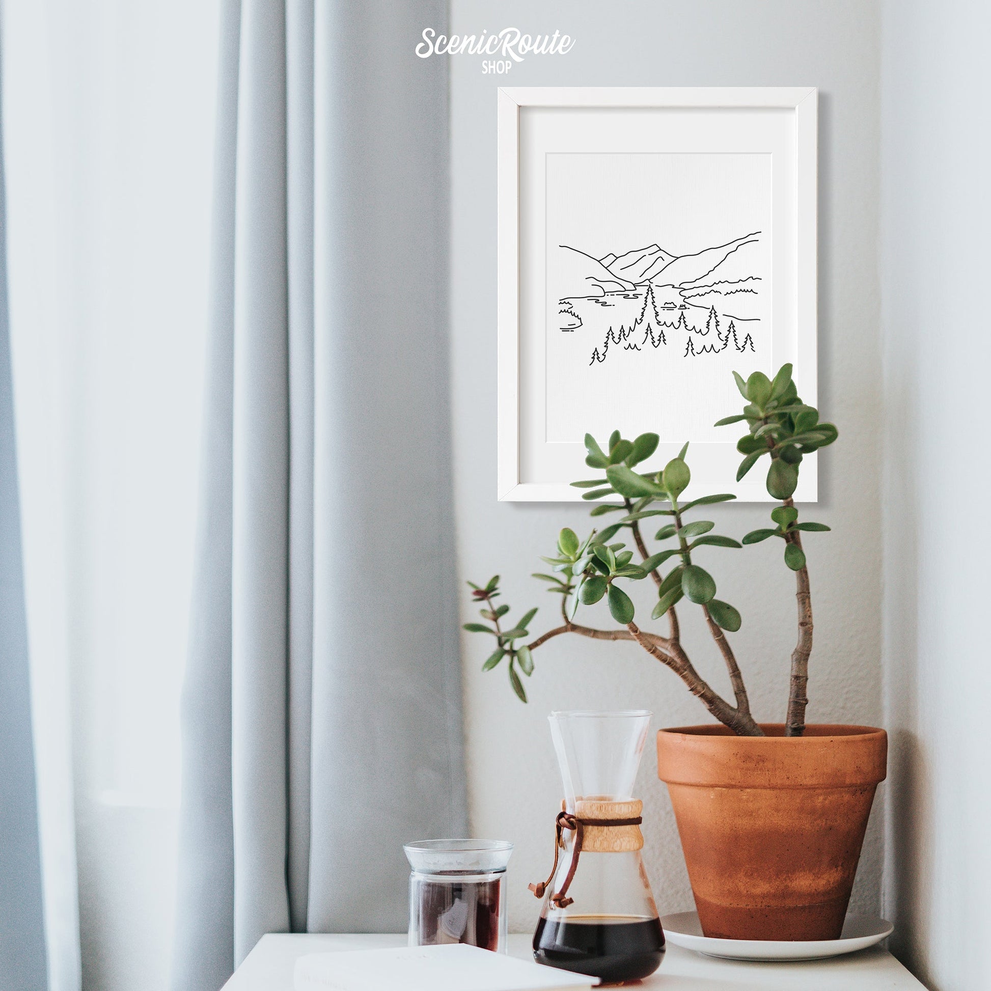 A framed line art drawing of North Cascades National Park on a wall above a table with a potted plant