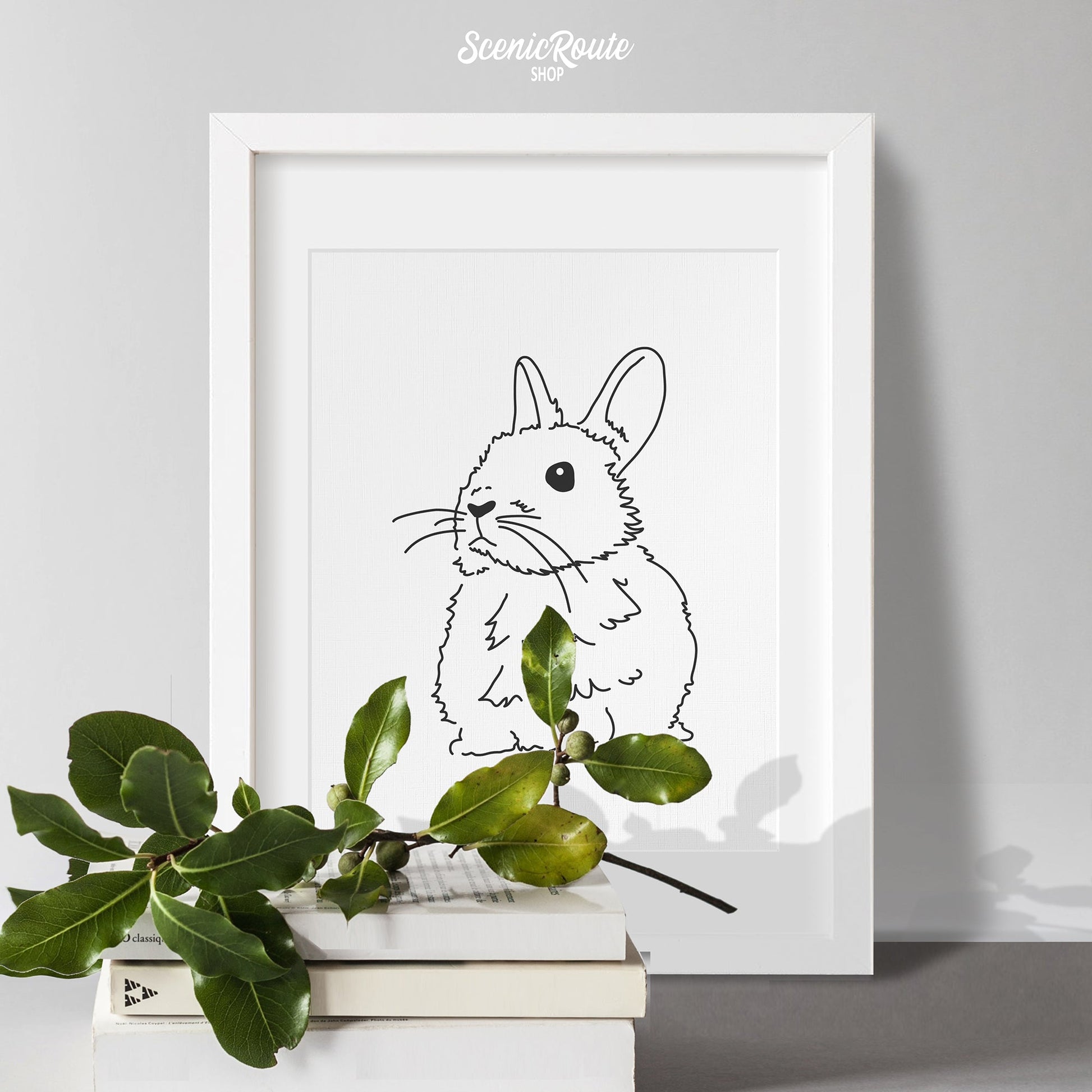A framed line art drawing of a Netherland Dwarf Rabbit on a table with books and a plant