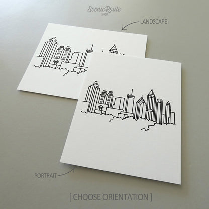 Two line art drawings of the Atlanta Skyline on white linen paper with a gray background.  The pieces are shown in portrait and landscape orientation for the available art print options.