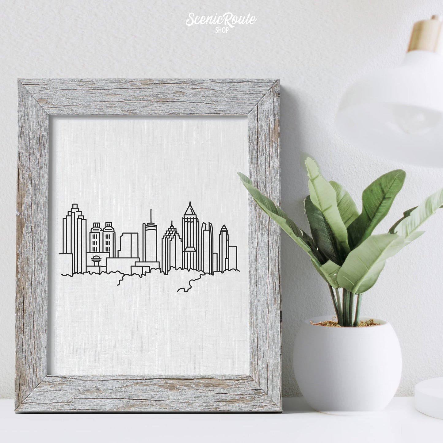 A framed line art drawing of the Atlanta Skyline on a desk with a lamp and plant