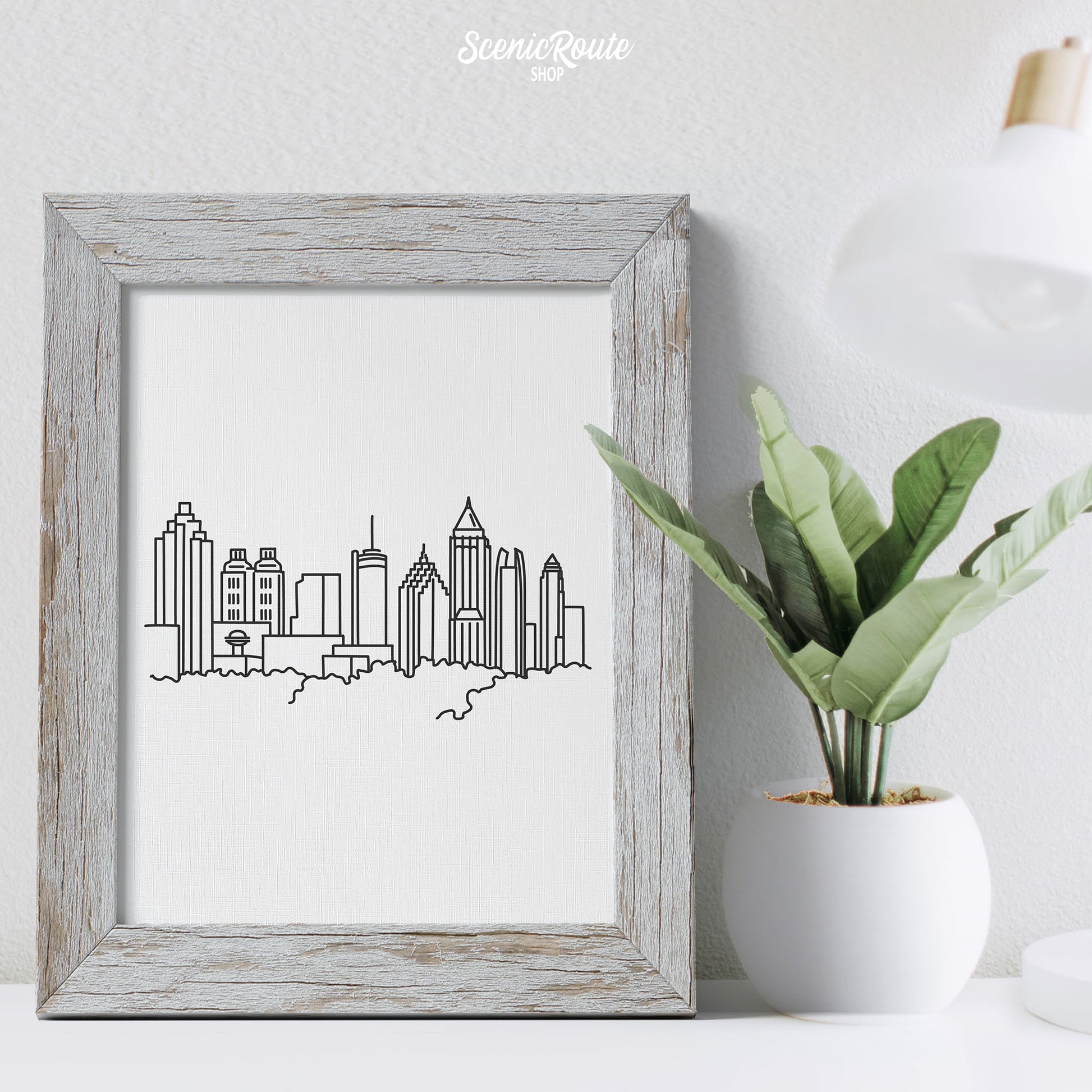 A framed line art drawing of the Atlanta Skyline on a desk with a lamp and plant