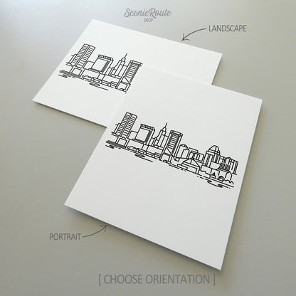 Two line art drawings of the Baltimore Skyline on white linen paper with a gray background.  The pieces are shown in portrait and landscape orientation for the available art print options.