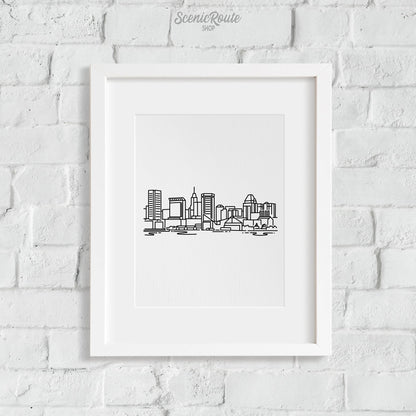 A framed line art drawing of the Baltimore Skyline hanging on a white brick wall
