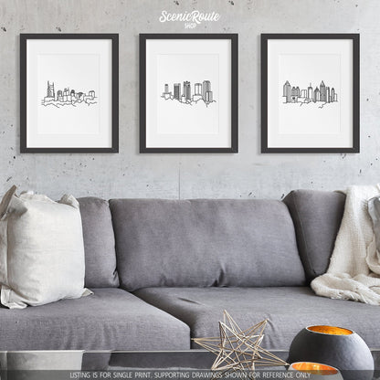 A group of three framed drawings on a white wall hanging above a couch with pillows and a blanket. The line art drawings include the Nashville Skyline, Birmingham Skyline, and Atlanta Skyline