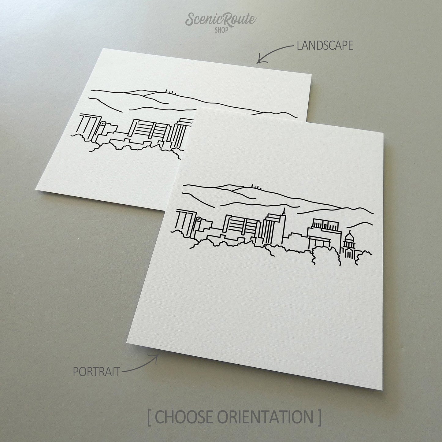 Two line art drawings of the Boise Skyline on white linen paper with a gray background.  The pieces are shown in portrait and landscape orientation for the available art print options.
