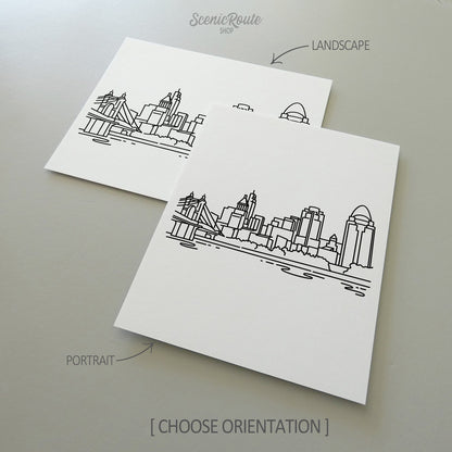 Two line art drawings of the Cincinnati Skyline on white linen paper with a gray background.  The pieces are shown in portrait and landscape orientation for the available art print options.