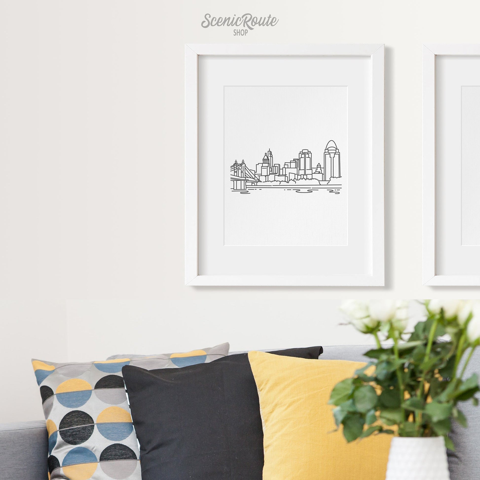 A framed line art drawing of the Cincinnati Skyline on a white wall above a couch with pillows