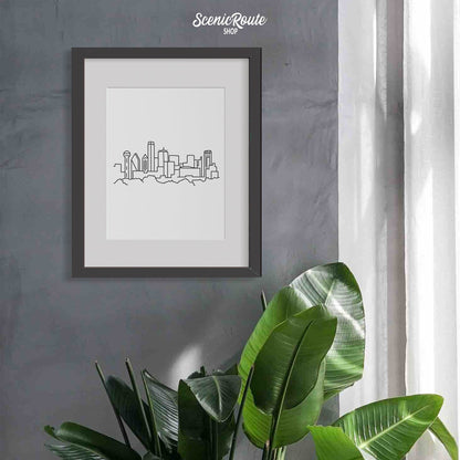A framed line art drawing of the Dallas Skyline hanging on a gray wall next to a window above a tropical plant