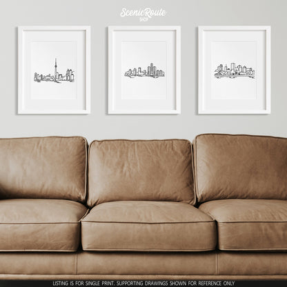 A group of three framed drawings on a wall above a leather couch. The line art drawings include the Toronto Skyline, Detroit Skyline, and Montreal Skyline