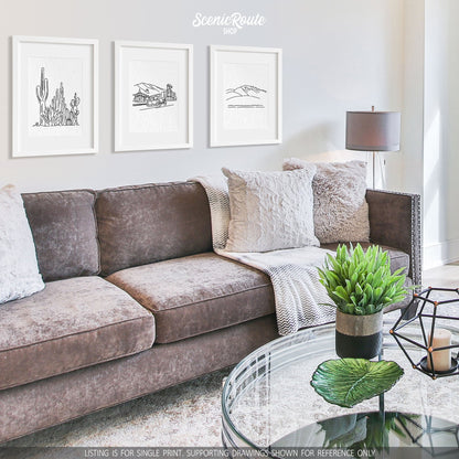 A group of three framed drawings on a white wall hanging above a couch with pillows and a blanket. The line art drawings include a Cactus Garden, Flagstaff Skyline, and Humphreys Peak