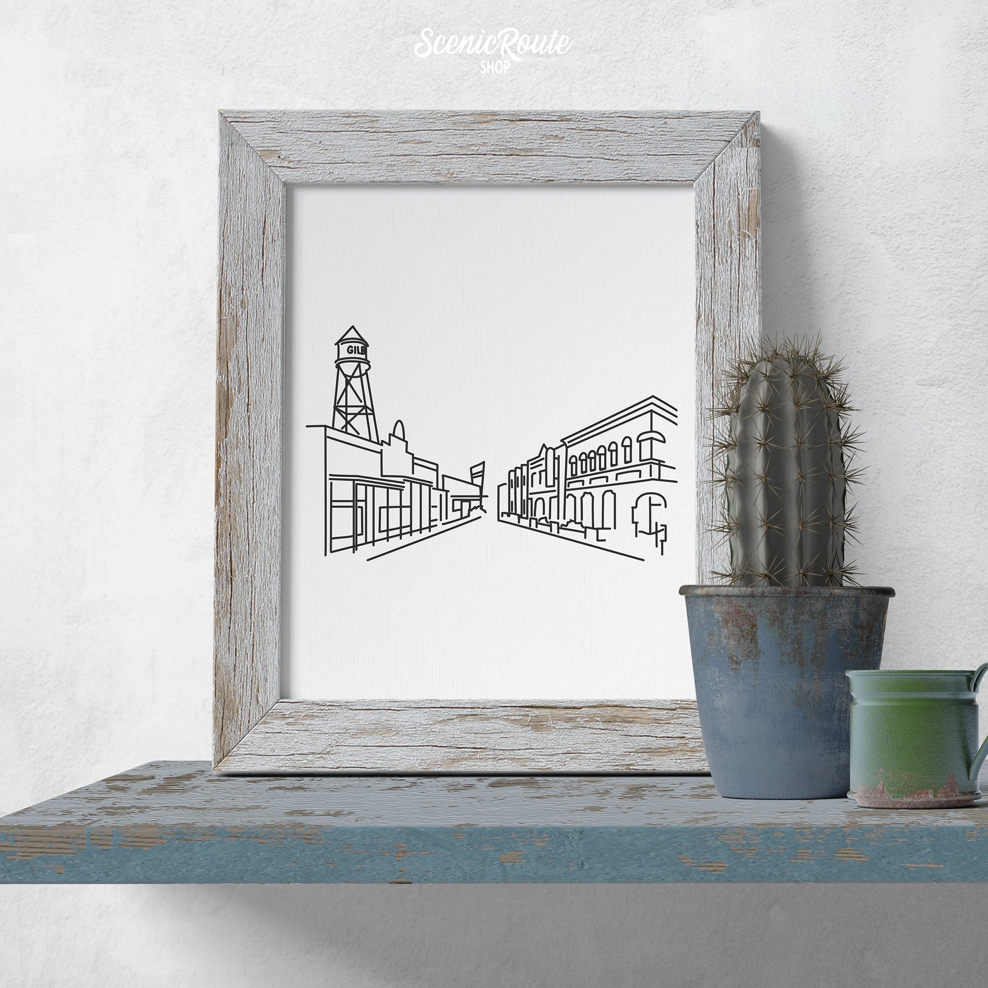 A framed line art drawing of the Gilbert Skyline on a blue shelf with a potted cactus
