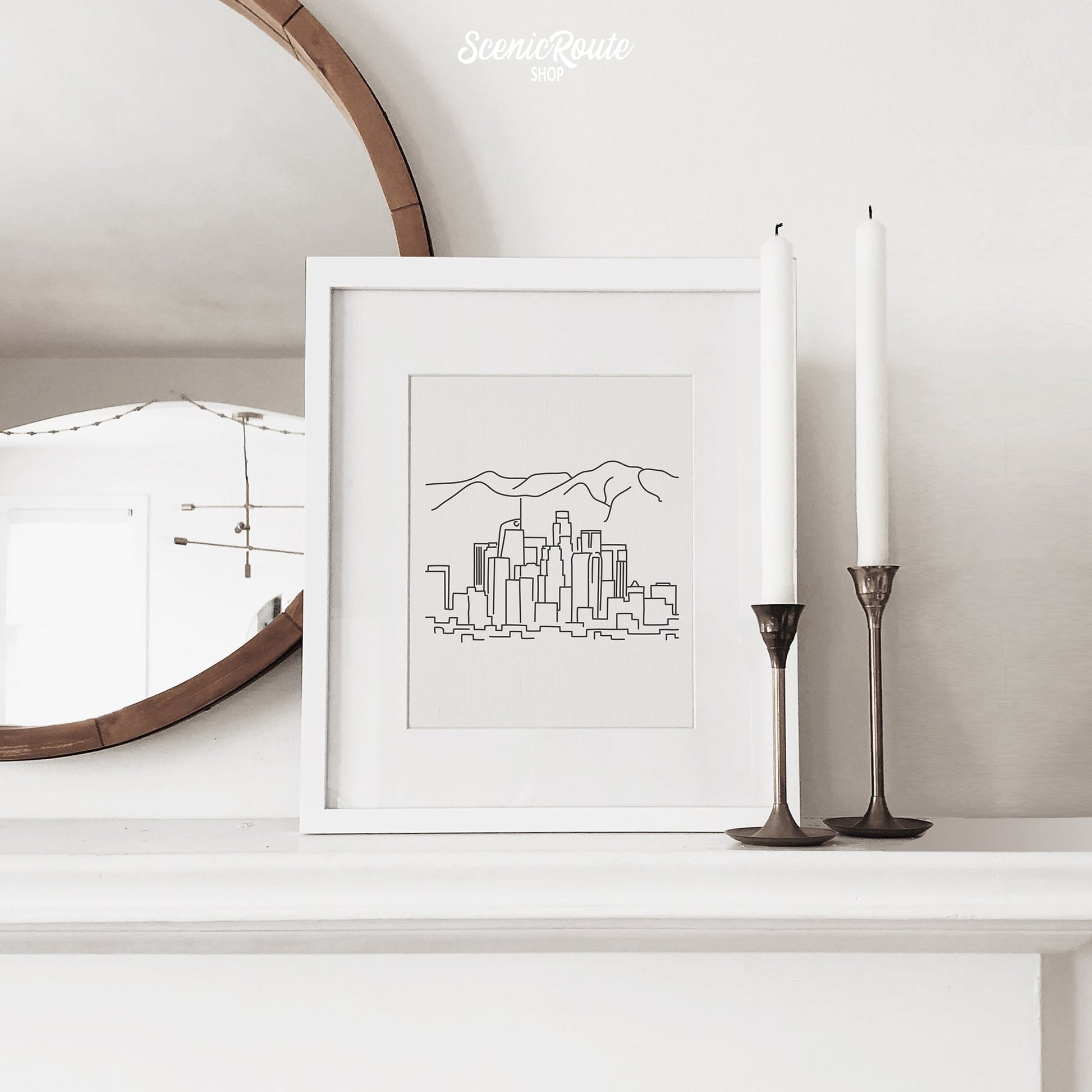 A framed line art drawing of the Los Angeles Skyline on a fireplace mantle with candles and a mirror