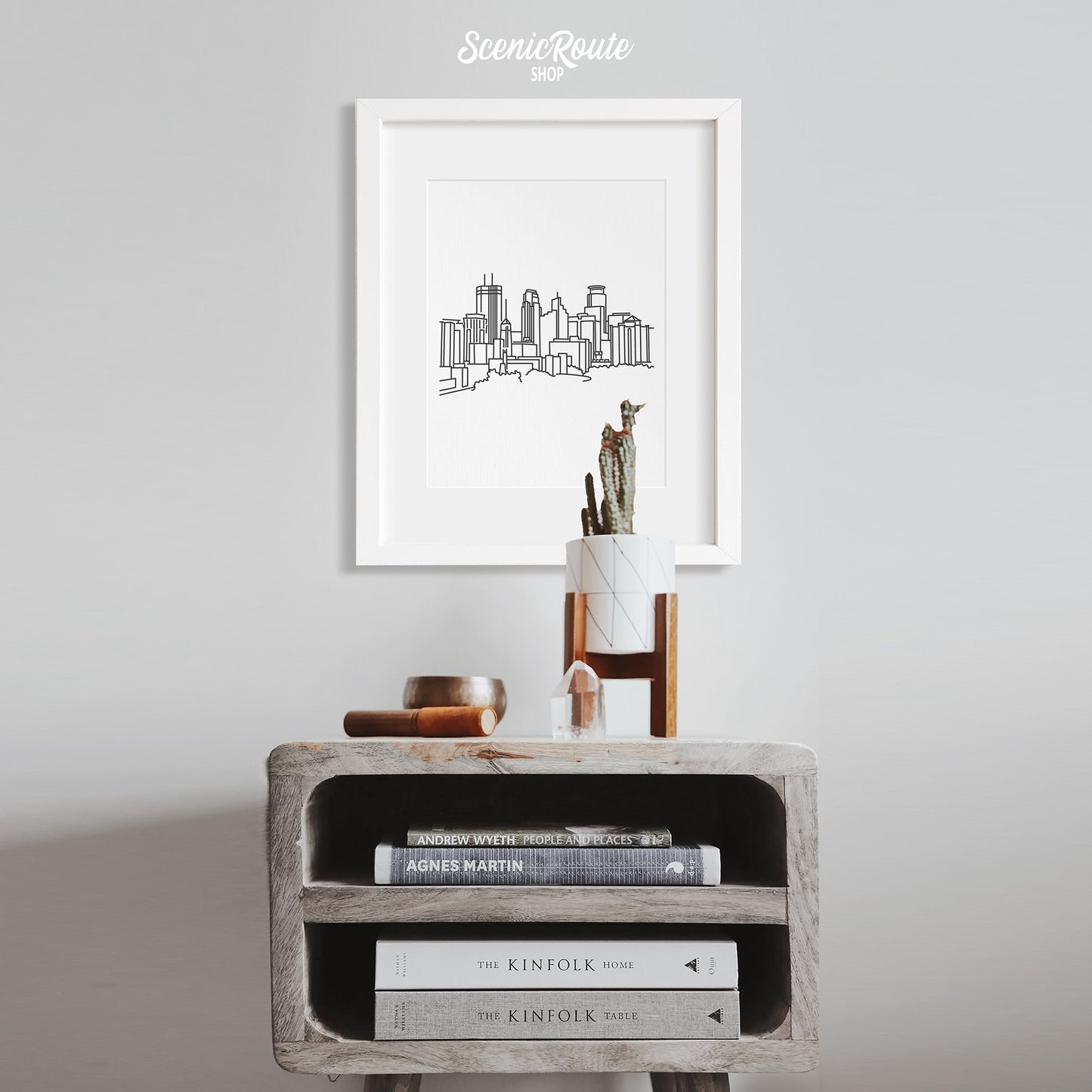 A framed line art drawing of the Minneapolis Skyline above a side table with a potted cactus
