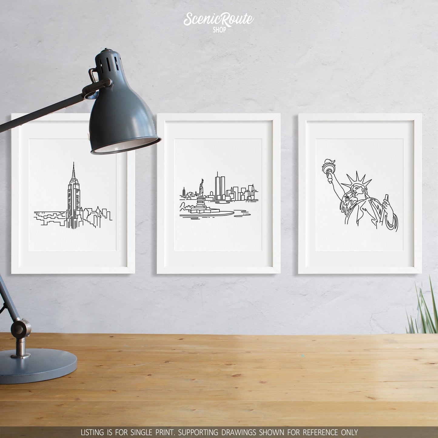 A group of three framed drawings on a wall above a desk with a lamp. The line art drawings include the Empire State Building, New York City Skyline, and Statue of Liberty