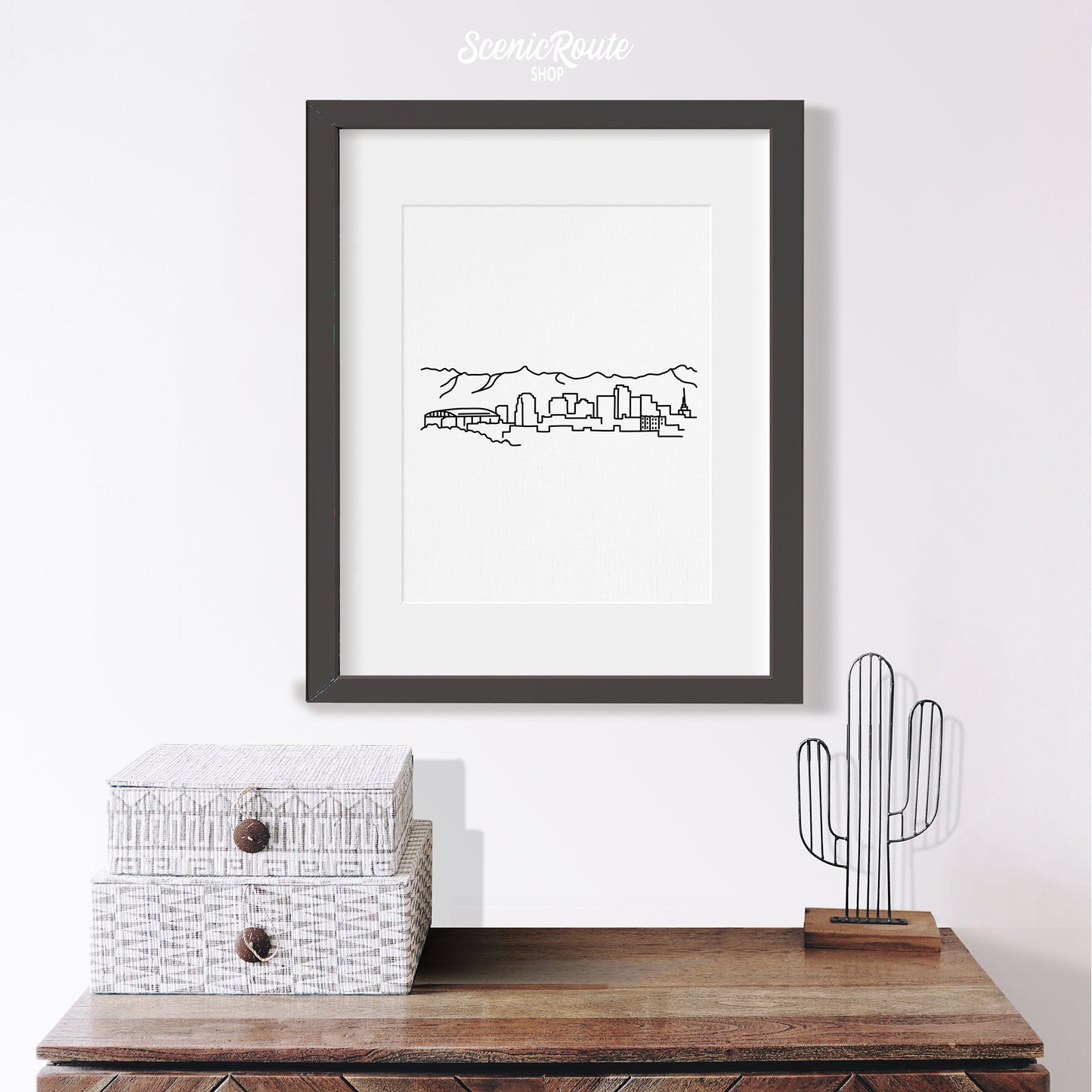 A framed line art drawing of the Phoenix Skyline hanging above a table with saguaro figurine