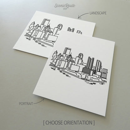 Two line art drawings of the Pittsburgh Skyline on white linen paper with a gray background.  The pieces are shown in portrait and landscape orientation for the available art print options.