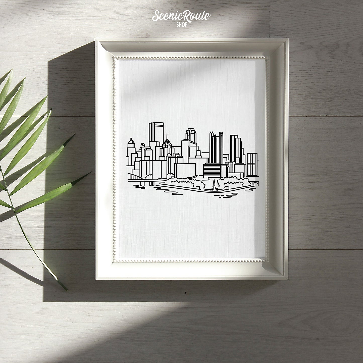 A framed line art drawing of the Pittsburgh Skyline laying on a wood table with a palm frond