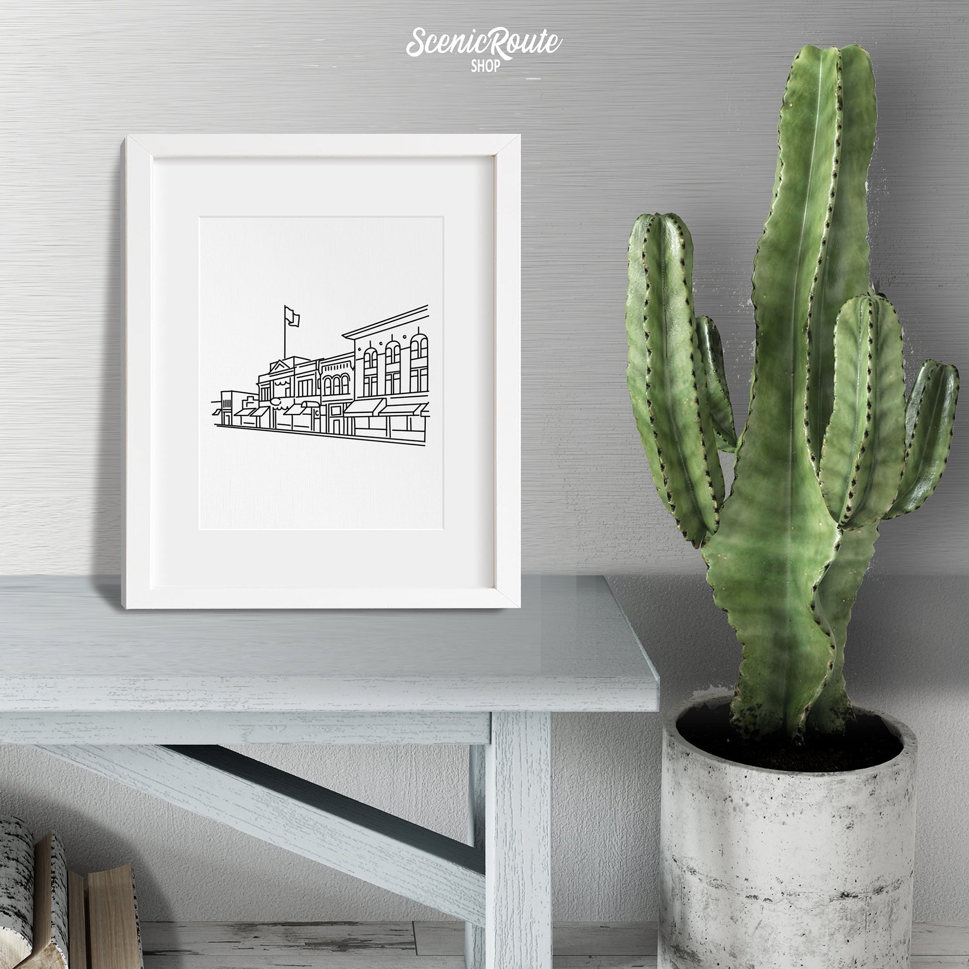 A framed line art drawing of the Prescott Skyline sitting on a wood bench next to a potted cactus