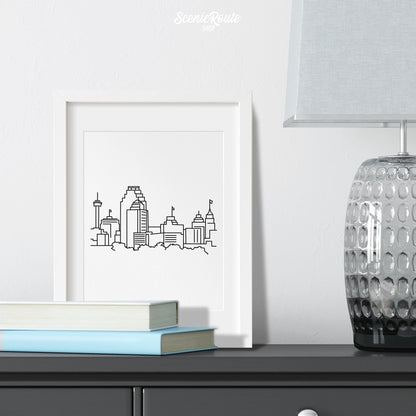 A framed line art drawing of the San Antonio Skyline on a dresser with books and a lamp
