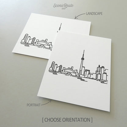 Two line art drawings of the Toronto Skyline on white linen paper with a gray background.  The pieces are shown in portrait and landscape orientation for the available art print options.