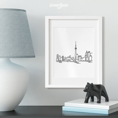 A framed line art drawing of the Toronto Skyline above a table with a lamp and figurine