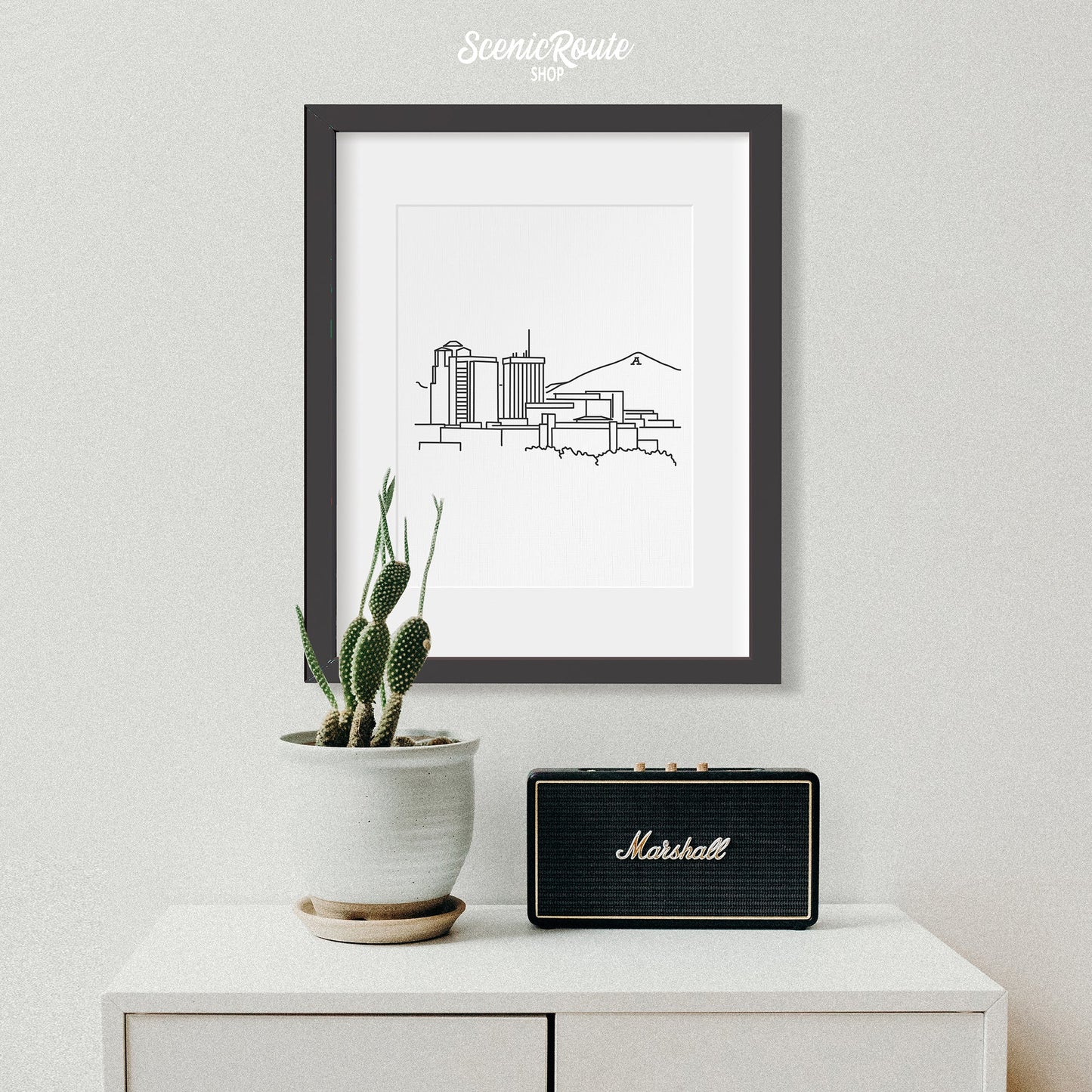 A framed line art drawing of the  Tucson Skyline above a side table with a potted cactus